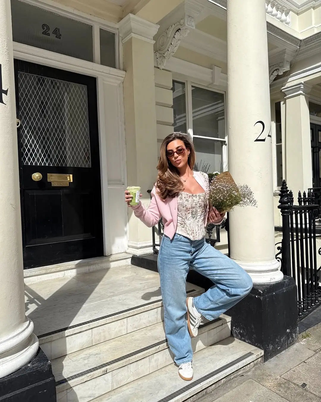 cream::Woman standing on steps in front of a building with columns, holding a drink and flowers.