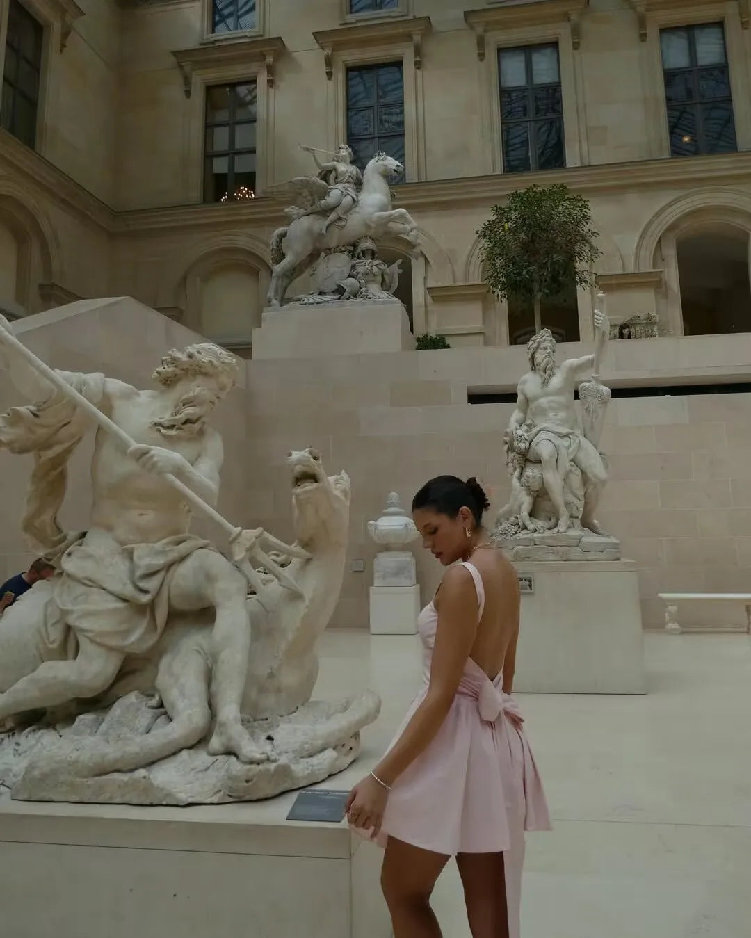 pink::Woman in a pink dress standing in front of classical statues in an outdoor setting.