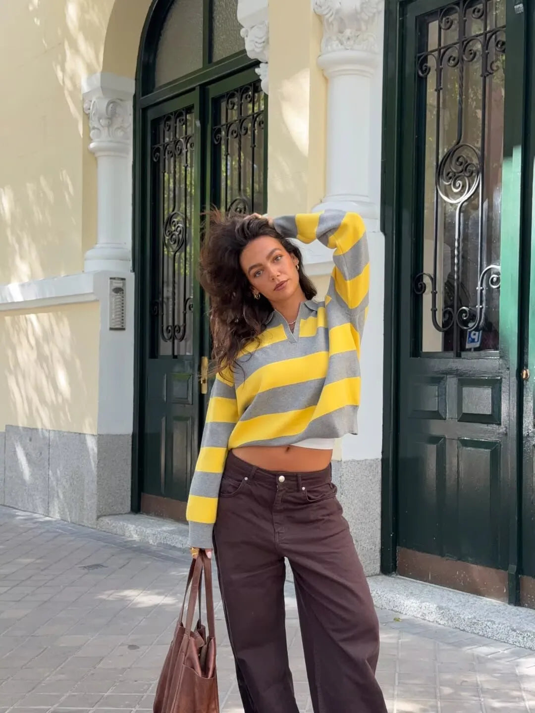 stripped::Woman wearing a yellow and gray striped sweater and brown pants standing in front of a building entrance.