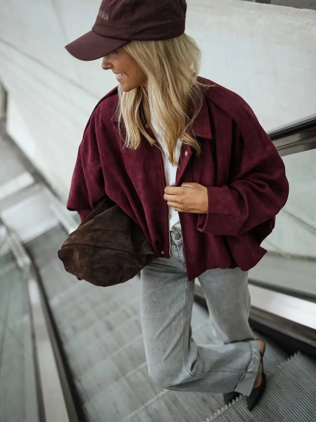 burgundy::Woman wearing a burgundy jacket and cap, holding a brown bag, on an escalator.