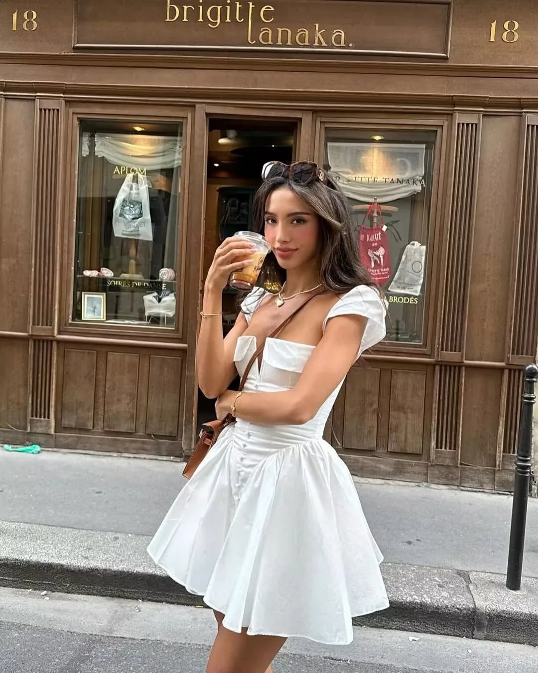 White:: Woman in a white dress standing on a street in front of a building with 'brigitte lanaka' sign.