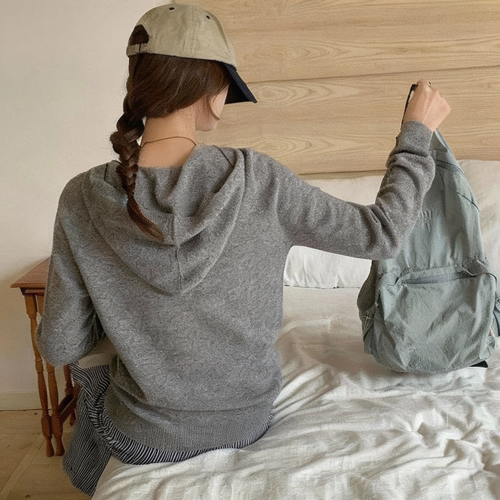 Light Grey:: Person wearing a gray hoodie and cap, sitting on a bed with a backpack.