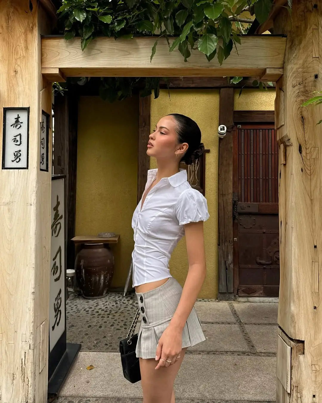 allvariant::Woman in a white blouse and gray skirt standing in front of a traditional wooden building with yellow walls.
