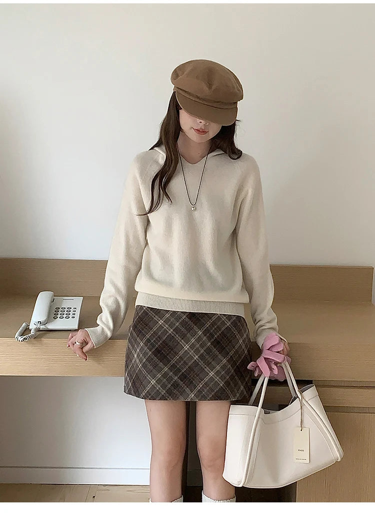 Cream:: Woman in beige sweater and plaid skirt holding a white handbag indoors.