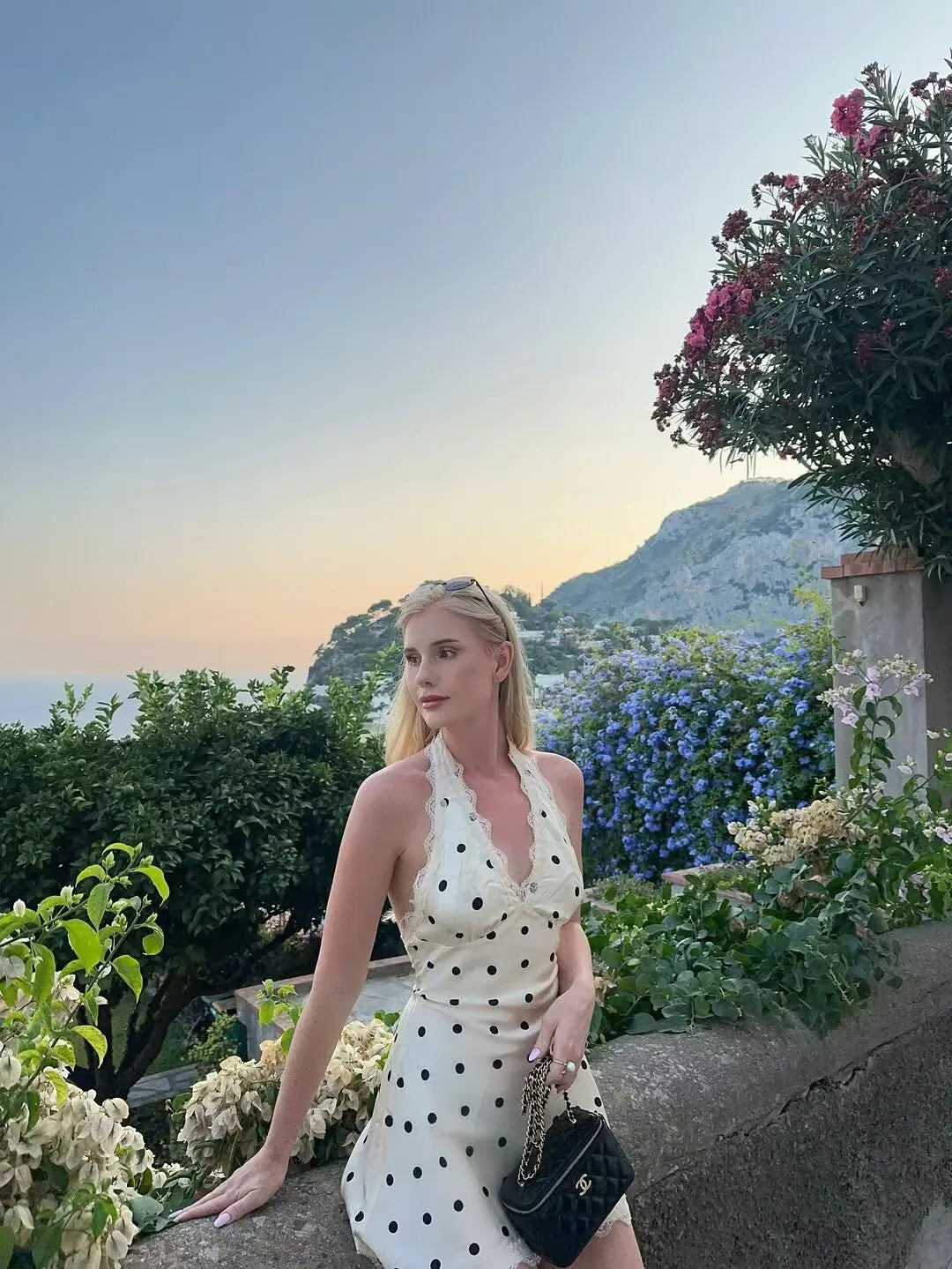 White::Woman in a white polka dot dress standing in a scenic outdoor setting with mountains and flowers.