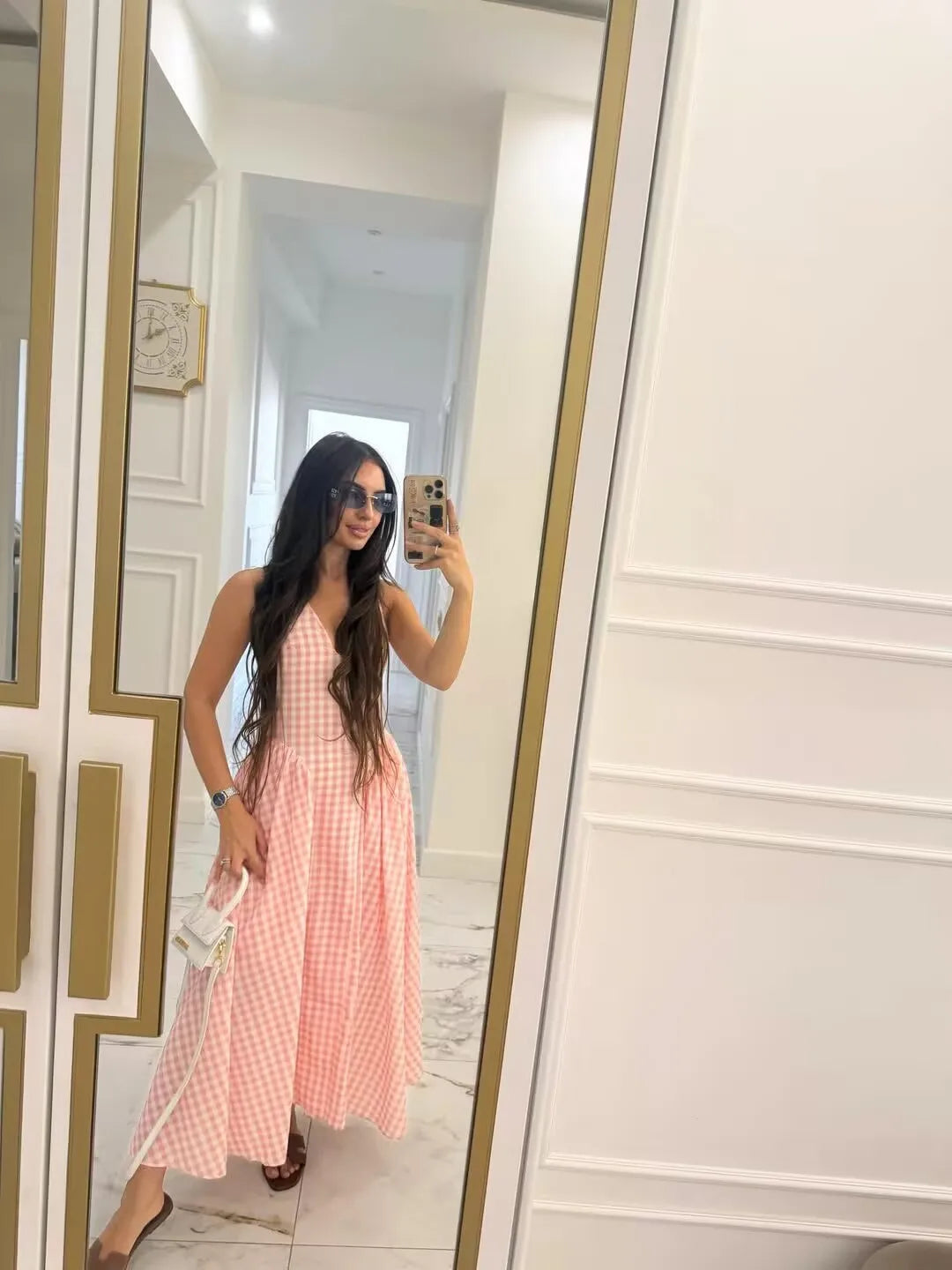 pink::Woman taking a mirror selfie wearing a pink checkered dress in a room with white walls and gold trim.