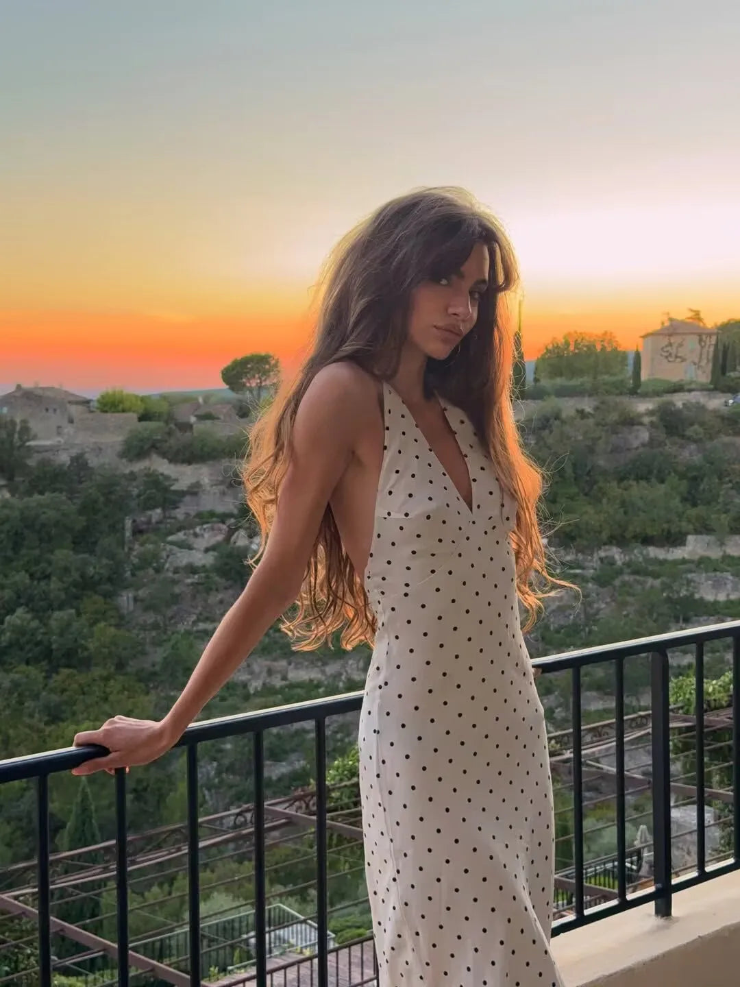 white::Woman in a polka dot dress standing on a balcony with a scenic view at sunset.