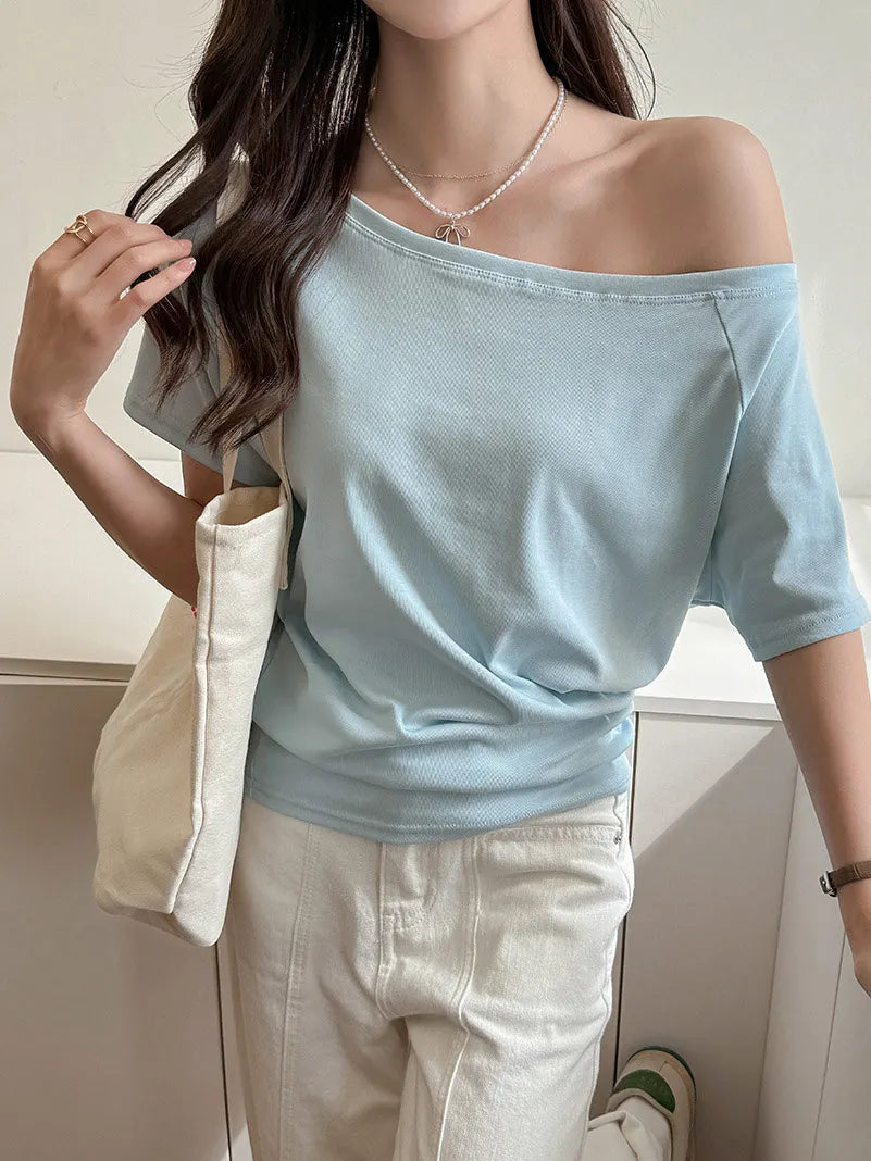allvariants:: Woman wearing a light blue off-shoulder top and beige pants, holding a beige bag indoors.