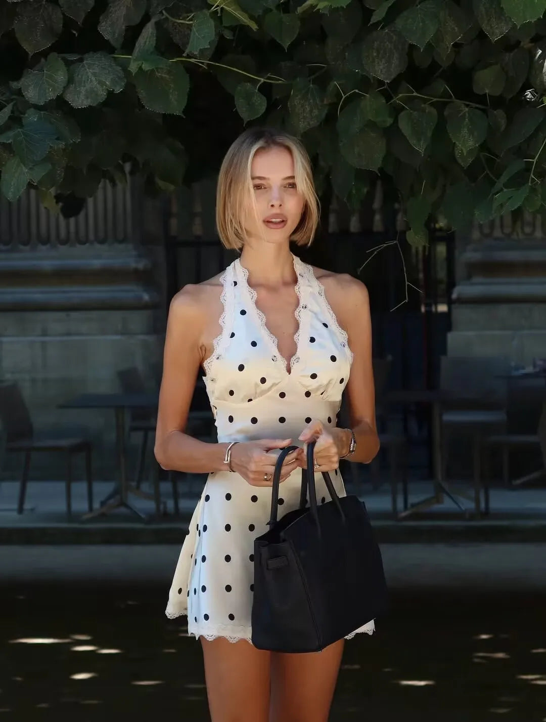 white::Woman in a polka dot dress holding a black handbag outdoors.