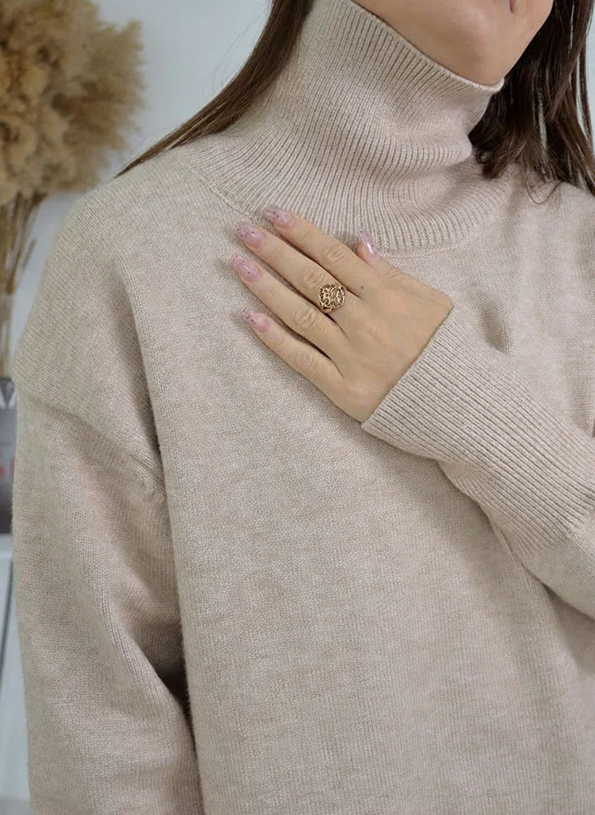 allvariants:: Person wearing a beige turtleneck sweater with a decorative ring on a blurred indoor background