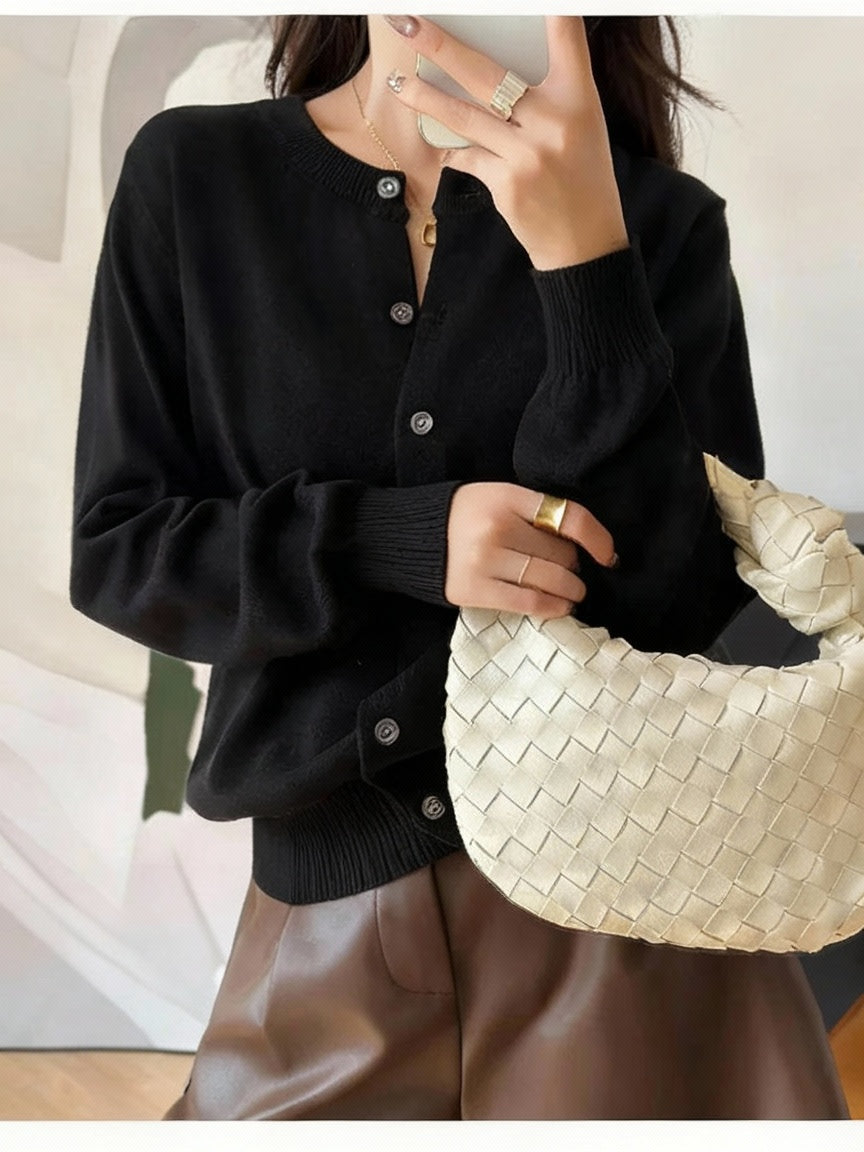 Black:: Person wearing a black cardigan holding a woven beige handbag indoors.