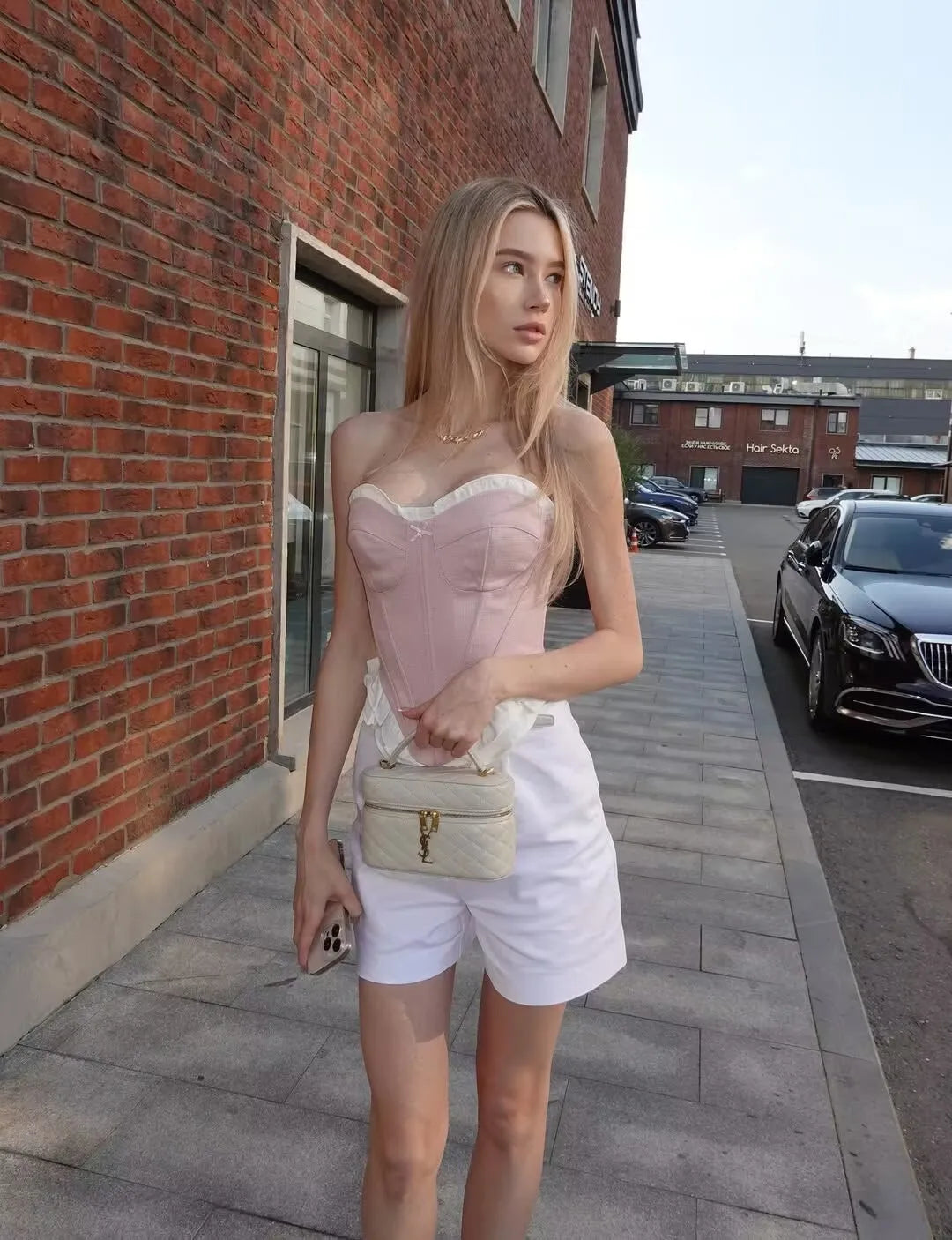 pink::Woman in a pink top and white shorts standing on a sidewalk with a brick building and parked cars in the background.
