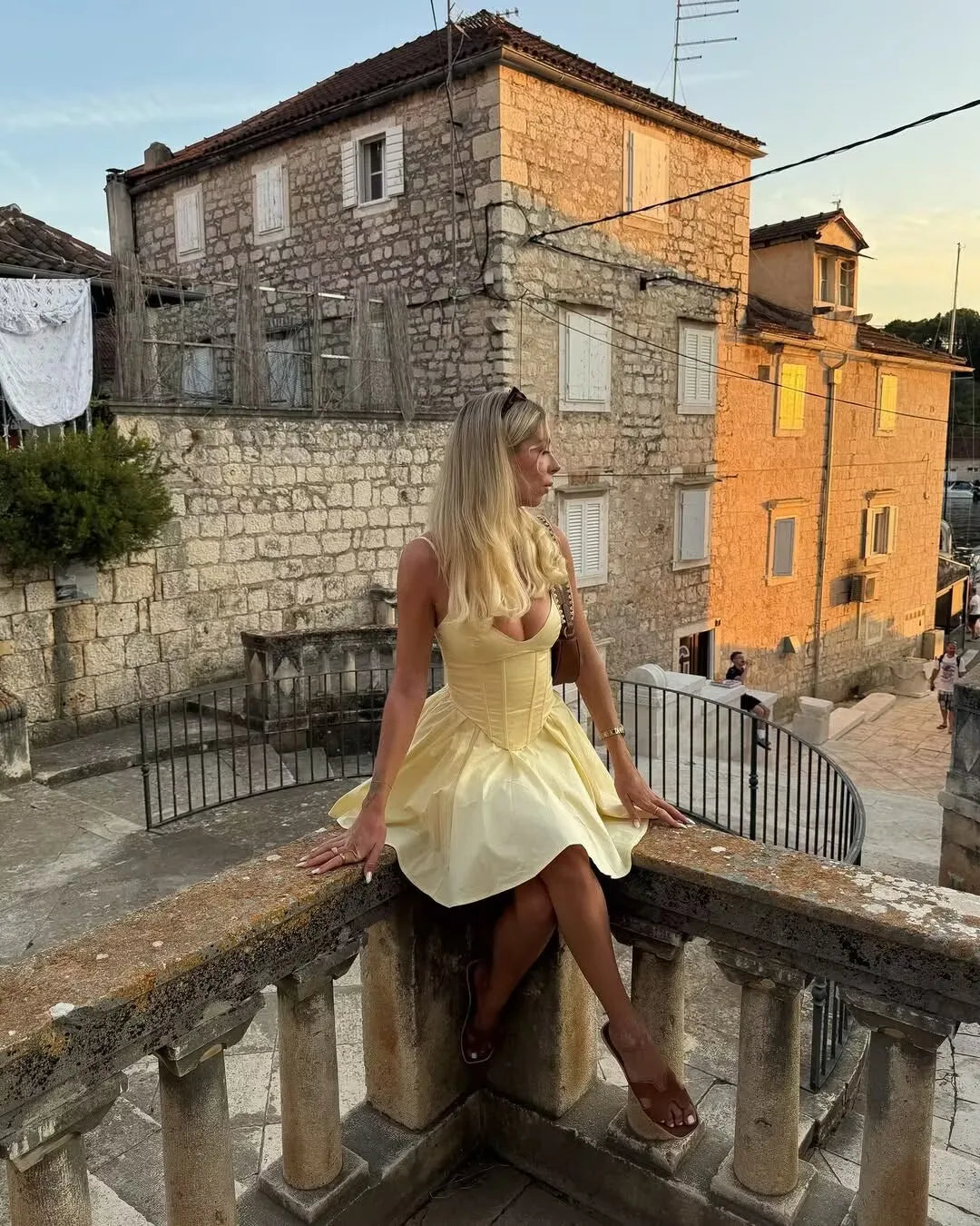 allvariant::Woman in a yellow dress sitting on a stone railing in a sunlit street with old buildings.