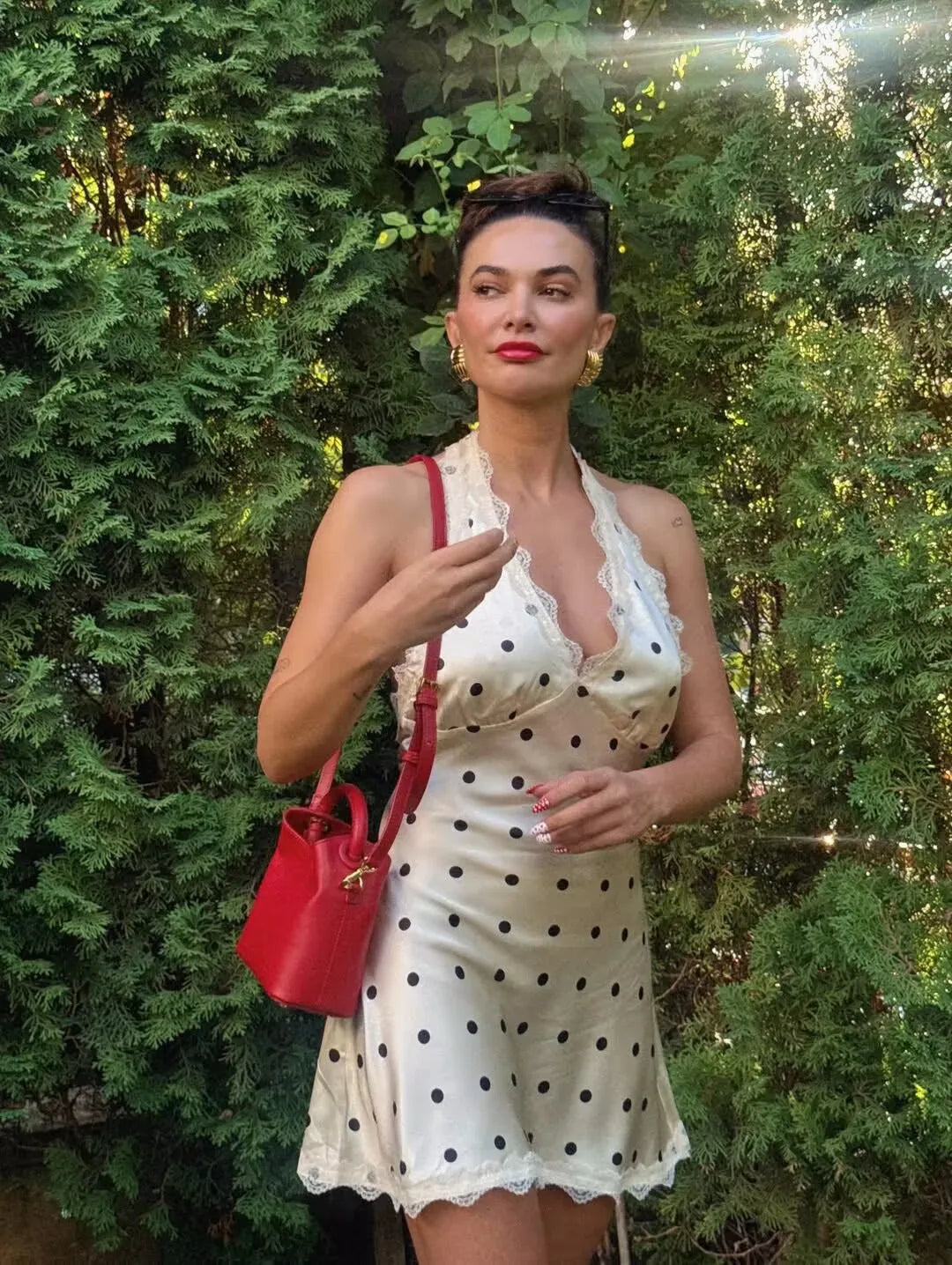 white::Woman in a white polka dot dress holding a red handbag in front of green foliage
