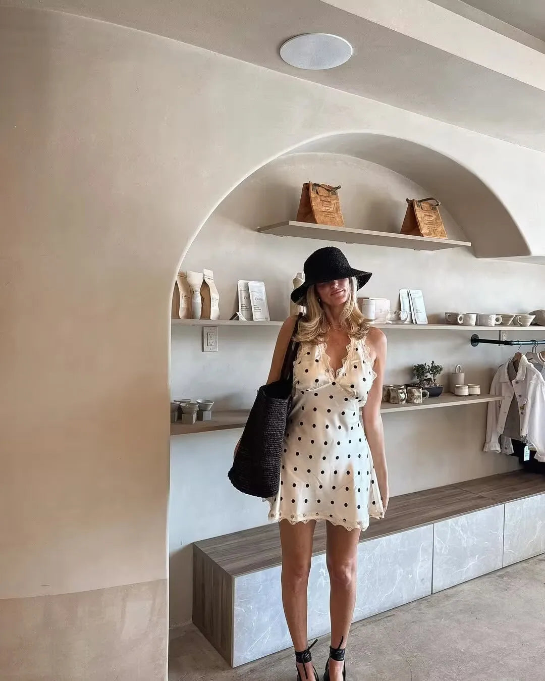 White::Woman in a polka dot dress and black hat standing in a modern interior setting.