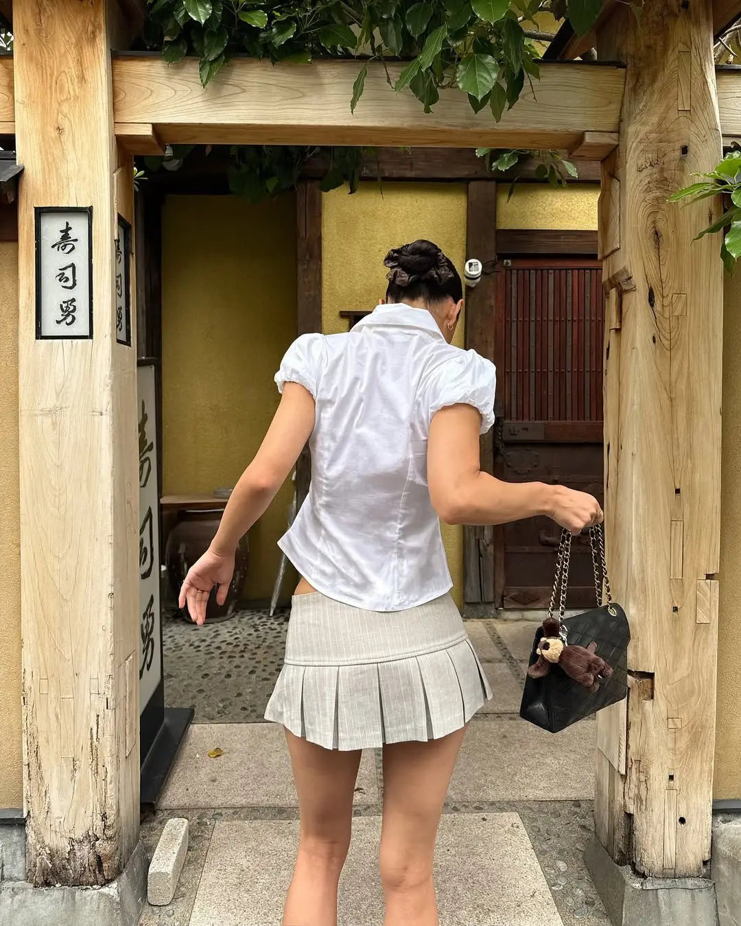 allvariant::Woman in a white shirt and pleated skirt walking through a traditional Japanese doorway.