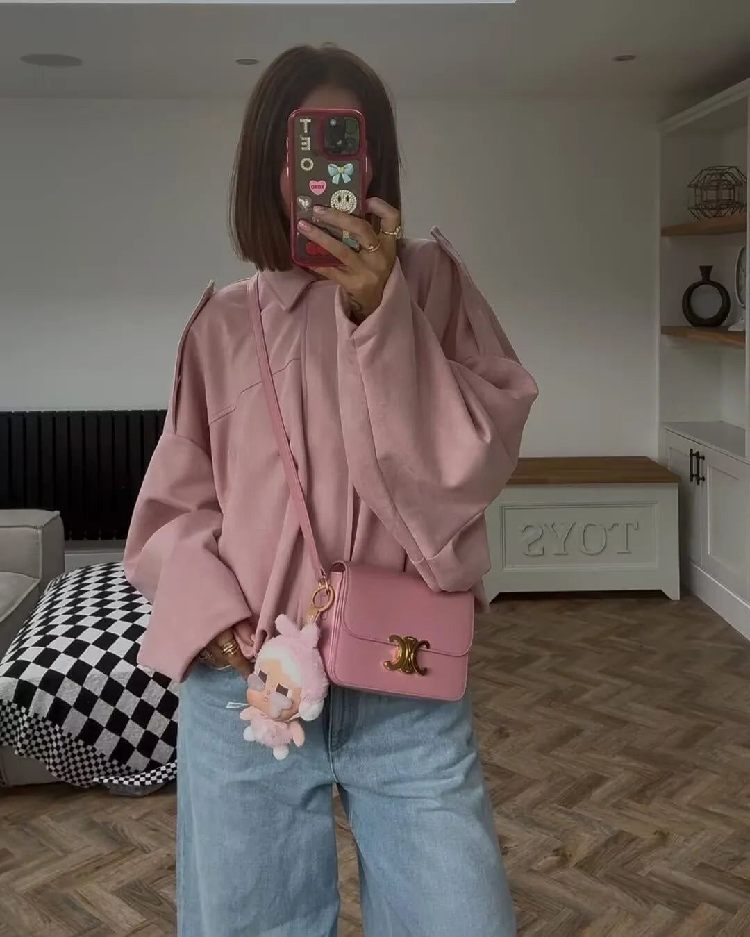 pink::Person wearing a pink jacket and holding a pink bag in a room with a checkered ottoman and white cabinet.