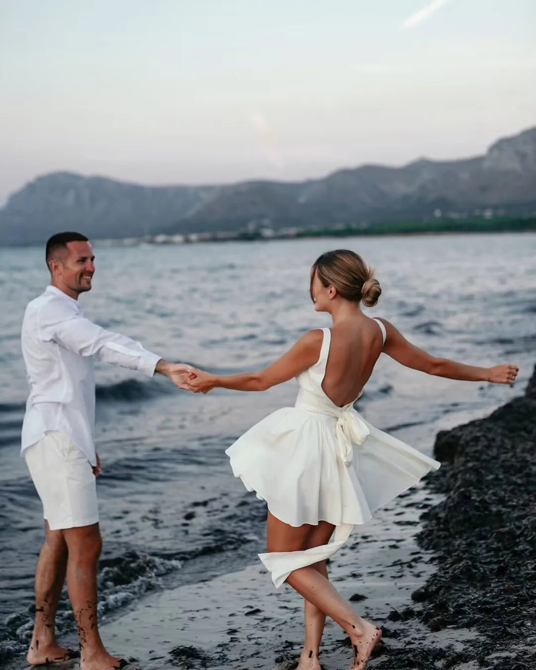 white::Couple in white outfits standing on a beach with mountains in the background